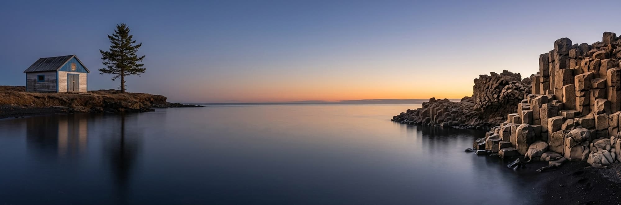 Casa solitaria junto a un lago en calma al atardecer, con rocas oscuras en la orilla.