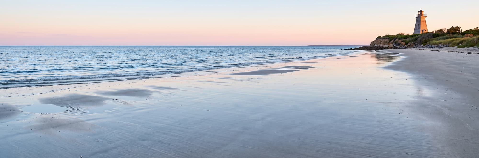 Playa de arena mojada al amanecer, con un faro a lo lejos y el mar en calma.